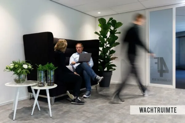 Twee mensen zitten in een moderne wachtkamer op de Baarnsche Dijk terwijl iemand voorbijloopt.